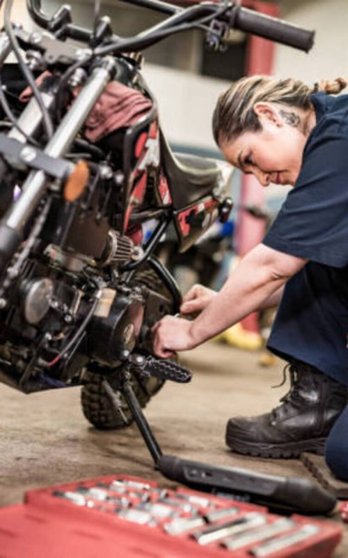 Mexican female mechanic in her shop. Mid adult woman with braided hair, wearing work clothes. Interior of car and motorcycle repair shop during day.
